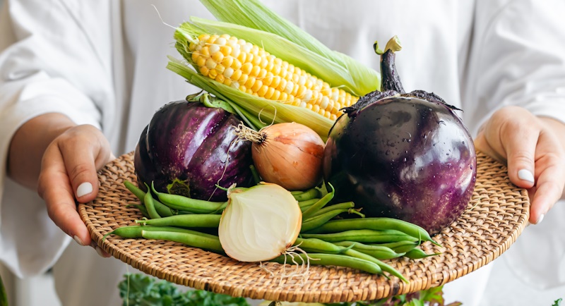 A woman holds a plate with eggplant, onion, corn and green beans, close-up.