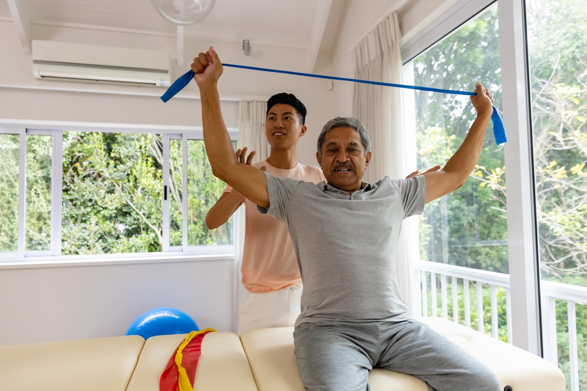 Happy diverse male physiotherapist advising and senior male patient using exercise band. Physiotherapy, healthcare, wellbeing, and senior lifestyle, unaltered.